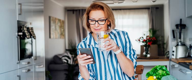 Middle-aged woman in her kitchen, drinking a glass of water while looking at her cell phone.