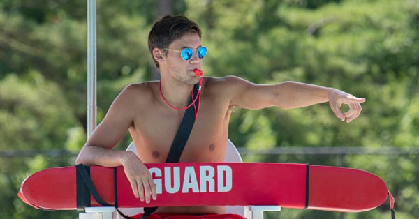 Lifeguard in chair with sunglasses and whistle in his mouth pointing at someone.
