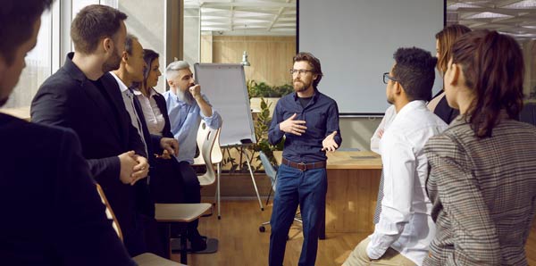 Young man talking with attendees – casually gathered around him – in a classroom.